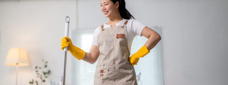 Beautiful asian woman wearing apron and gloves holding a cleaning mop posing in the bedroom Smiling asian female cleaning, wearing protective gear, holding mop in residential bedroom setting