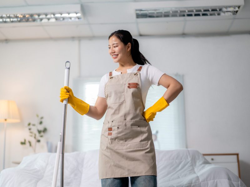 Smiling asian female cleaning, wearing protective gear, holding mop in residential bedroom setting