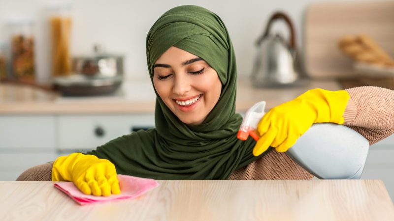 Closeup portrait of happy islamic housewife in hijab and protecitve yellow rubber gloves cleaning dining table at kitchen. Positive maid keeping her home tidy, wiping surface with microfiber cloth