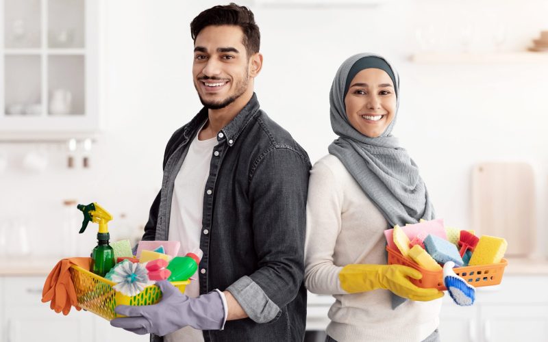Happy muslim family cleaning their new apartment, holding house-keeping supplies and standing back to back in white kitchen, smiling at camera, copy space. House-keeping, apartment cleaning concept