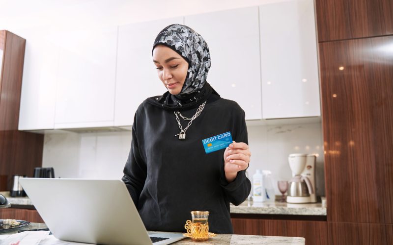 Cheerful young female in hijab is drinking tea in kitchen and doing online shopping with laptop