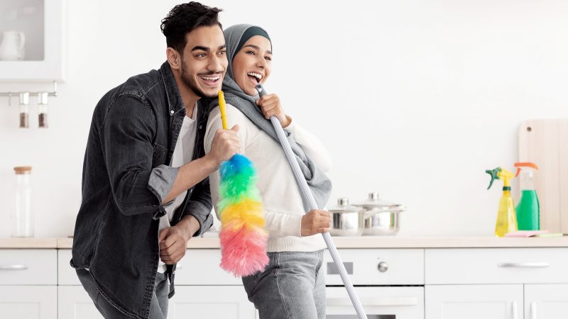 Beautiful joyful muslim young couple cleaning floor and having fun in kitchen together, full length photo, copy space. Funny lady in hijab and her singing boyfriend house-keeping their new house