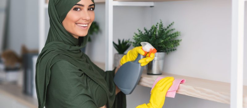Muslim young woman cleaning shelfs with detergent at home Hygiene And Cleanliness. Portrait of happy muslim lady in hijab cleaning bookshelf surface in living room, bedroom or office, wiping dust holding spray bottle using microfiber cloth, looking at camera