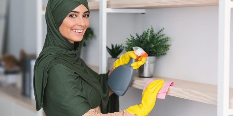Hygiene And Cleanliness. Portrait of happy muslim lady in hijab cleaning bookshelf surface in living room, bedroom or office, wiping dust holding spray bottle using microfiber cloth, looking at camera
