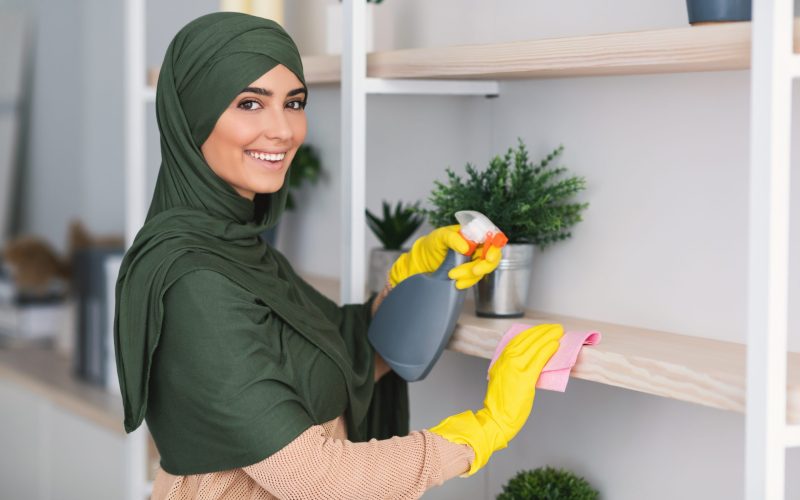 Muslim young woman cleaning shelfs with detergent at home Hygiene And Cleanliness. Portrait of happy muslim lady in hijab cleaning bookshelf surface in living room, bedroom or office, wiping dust holding spray bottle using microfiber cloth, looking at camera