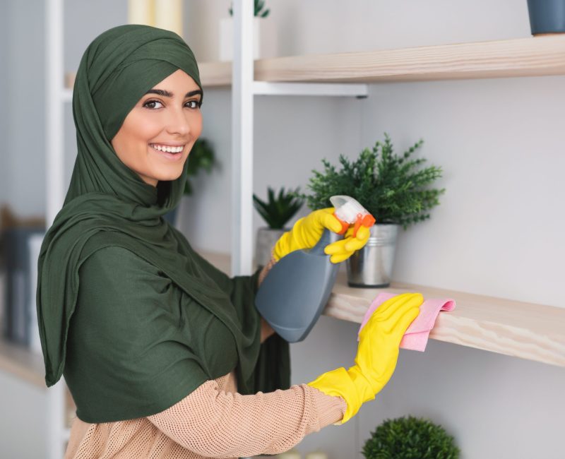 Muslim young woman cleaning shelfs with detergent at home Hygiene And Cleanliness. Portrait of happy muslim lady in hijab cleaning bookshelf surface in living room, bedroom or office, wiping dust holding spray bottle using microfiber cloth, looking at camera
