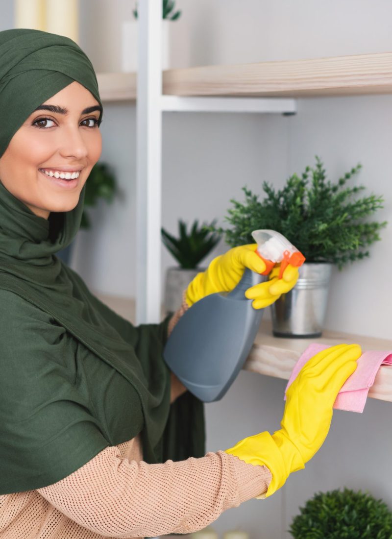 Hygiene And Cleanliness. Portrait of happy muslim lady in hijab cleaning bookshelf surface in living room, bedroom or office, wiping dust holding spray bottle using microfiber cloth, looking at camera