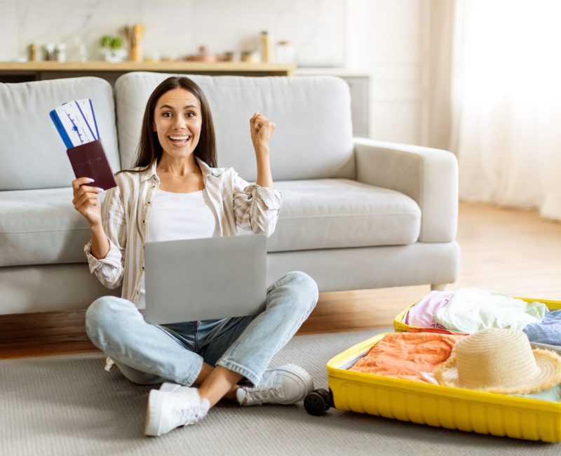 Time To Travel. Excited Young Woman With Laptop And Tickets Sitting Near Packed Suitcase, Happy European Female Holding Passport And Shaking Fist, Getting Ready For Journey, Copy Space